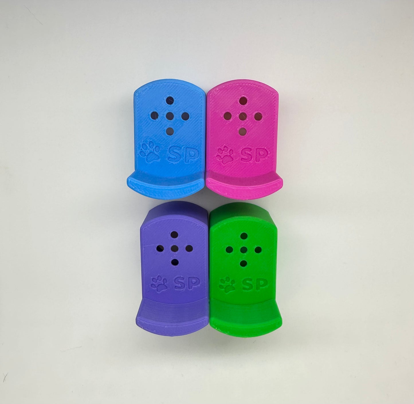 A set of four 3D-printed Sniffy Prints pairing extensions for K9 nosework. The extensions in the image are magnetic and designed to go over Sniffy Prints Tiny Hides. They are blue, pink, purple, and green. Each piece features a curved base to hold a treat, multiple small holes for odor to pass through, and an embossed 'SP' logo with a paw print.